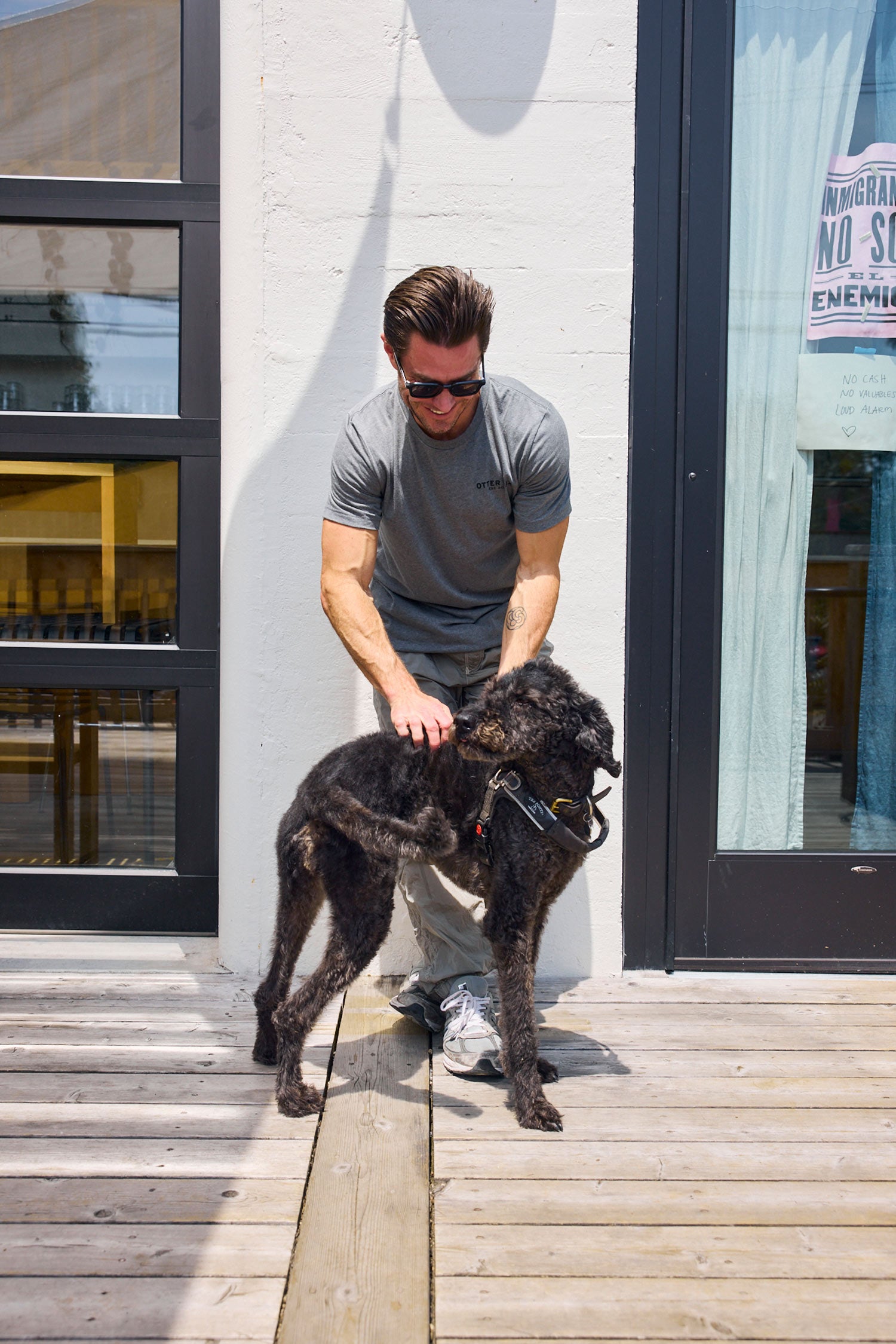Man with sunglasses and a dog on a wooden deck outside a building.