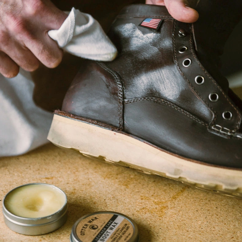 Person applying otter wax boot wax to a leather boot with a cloth, next to a tin of boot wax on a textured surface.