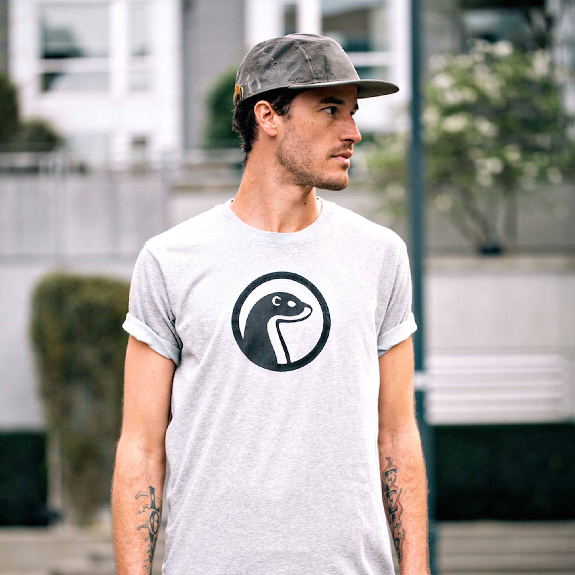 Man wearing a light gray Otter Wax t-shirt with the black otter logo and the word Apparel, standing outdoors in an urban setting.