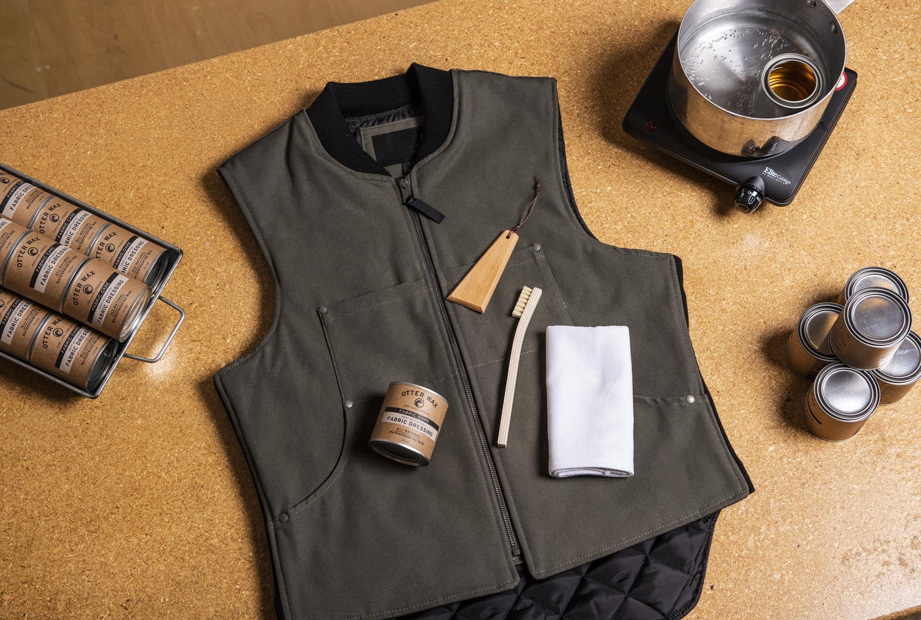 Waxed canvas vest on a cork surface with Otter Wax Fabric Dressing, brush, cloth, and wooden scraper beside a pot of melted wax and extra tins.