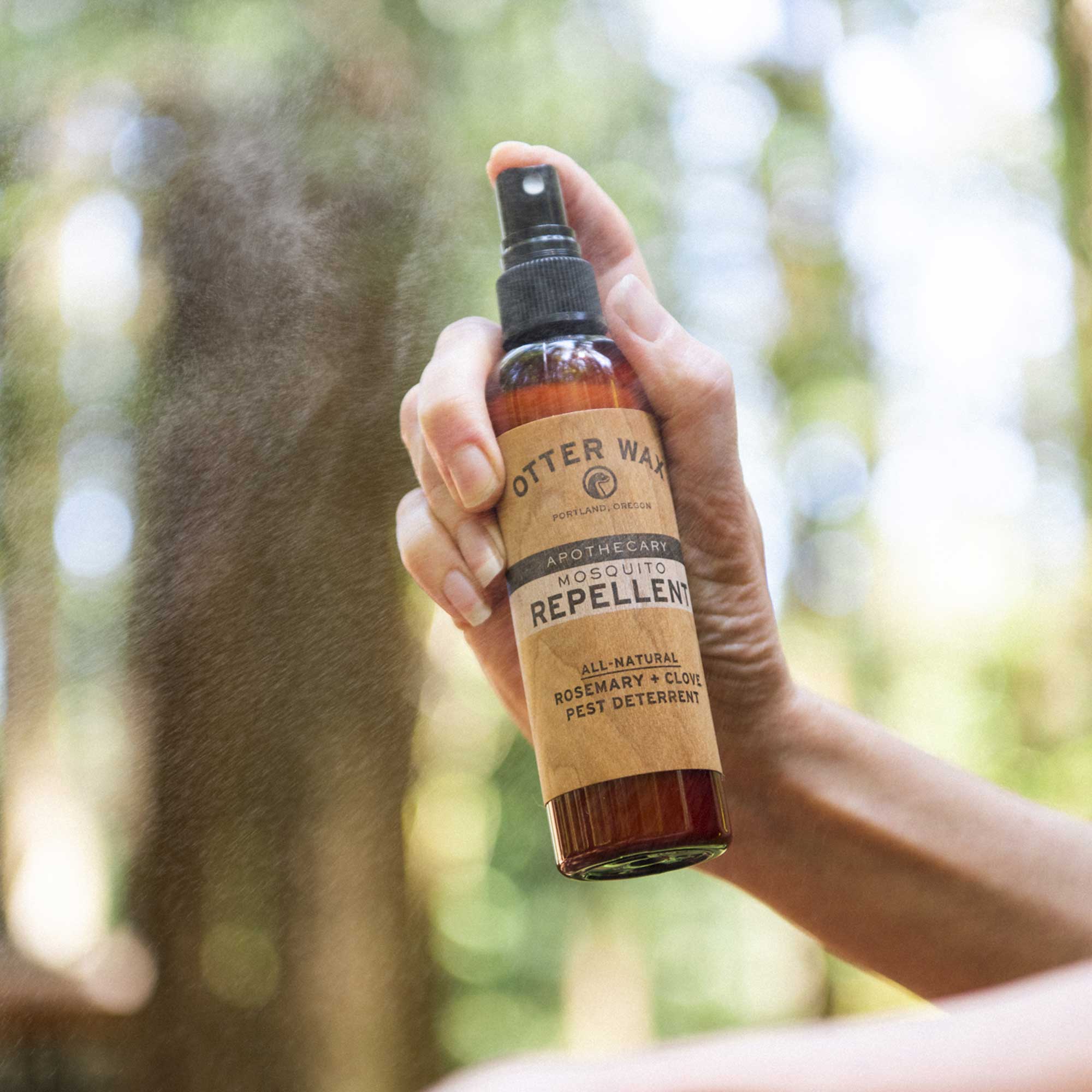 Hand spraying Otter Wax all-natural mosquito repellent bottle labeled with rosemary and clove ingredients, outdoors among blurred green trees.