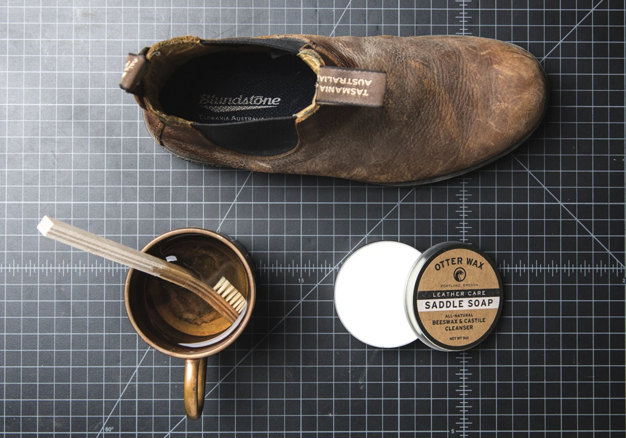 Otter Wax Saddle Soap tin open next to a worn Blundstone leather boot and natural fiber brush on a dark cutting mat