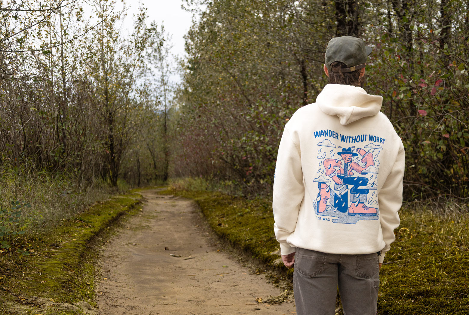 Person walking down a path in a forest wearing a hoodie featuring Otter Wax Wander Without Worry illustration.