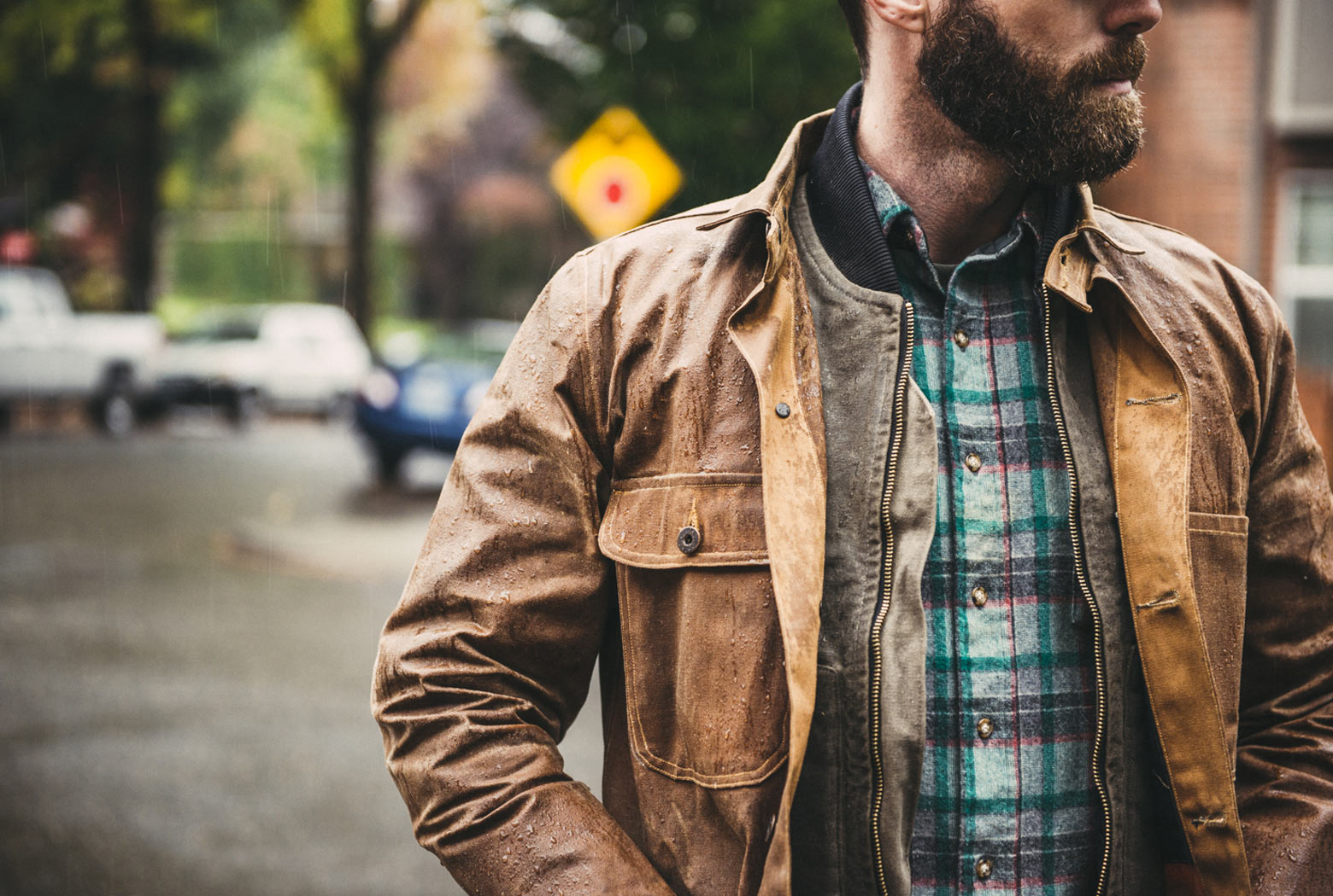 Close-up of a man wearing a rain-dappled waxed canvas jacket and plaid shirt, showing water beading on the fabric after waterproofing.
