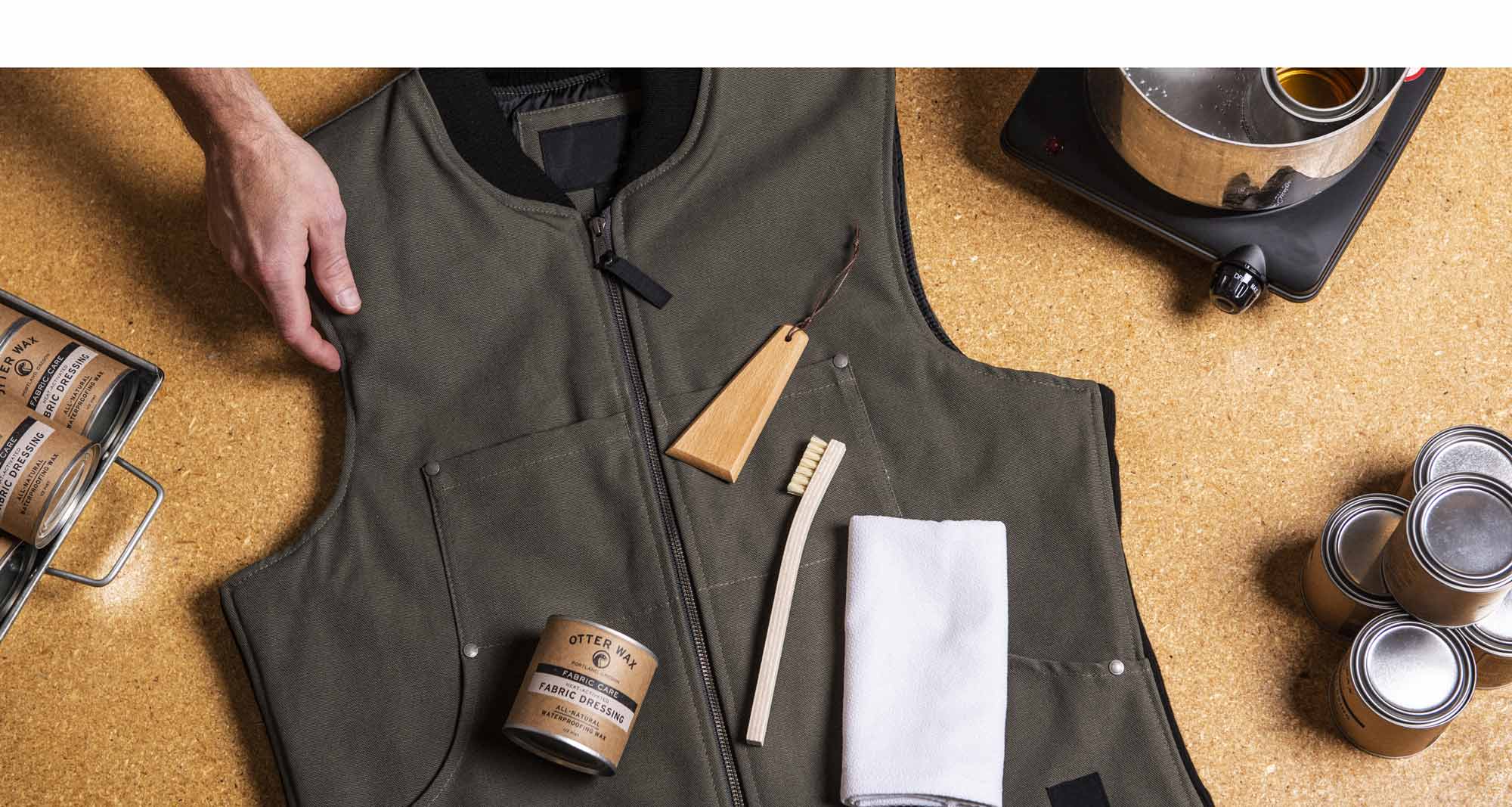 Person preparing to wax a green canvas vest with Otter Wax Fabric Dressing, brush, cloth, and wooden scraper on a cork work surface beside tins and a pot of melted wax.