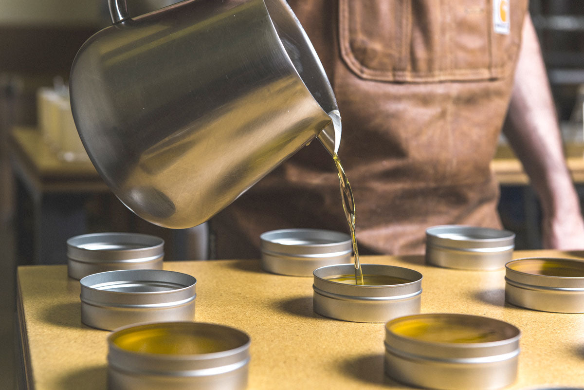 Person wearing a brown apron pouring melted wax from a metal pot into round tins on a workbench in the Otter Wax workshop.