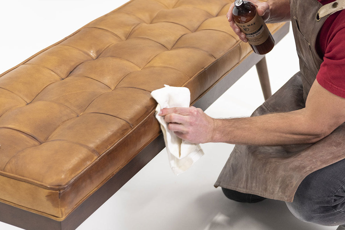 Person applying Otter Wax Leather Oil to a tan leather bench using a white cloth while wearing an apron.