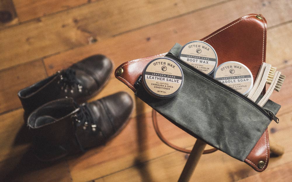 Otter Wax Leather Care Kit with tins of Leather Salve, Boot Wax, and Saddle Soap inside a waxed canvas pouch beside a pair of worn leather boots on a wooden floor.