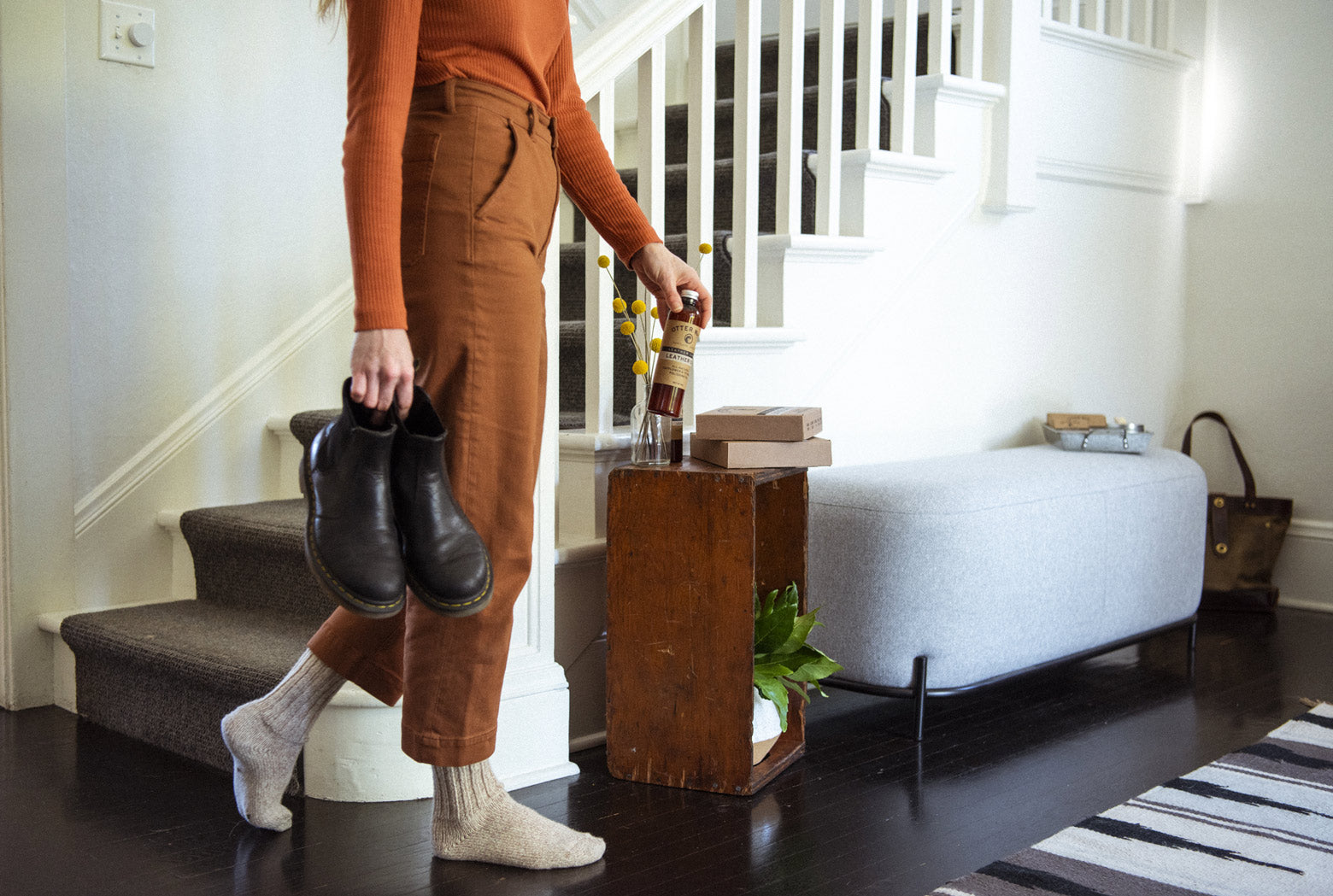 Person standing near a staircase holding black leather boots and a bottle of Otter Wax Leather Cleaner, with a wooden crate and small bench in a bright entryway.