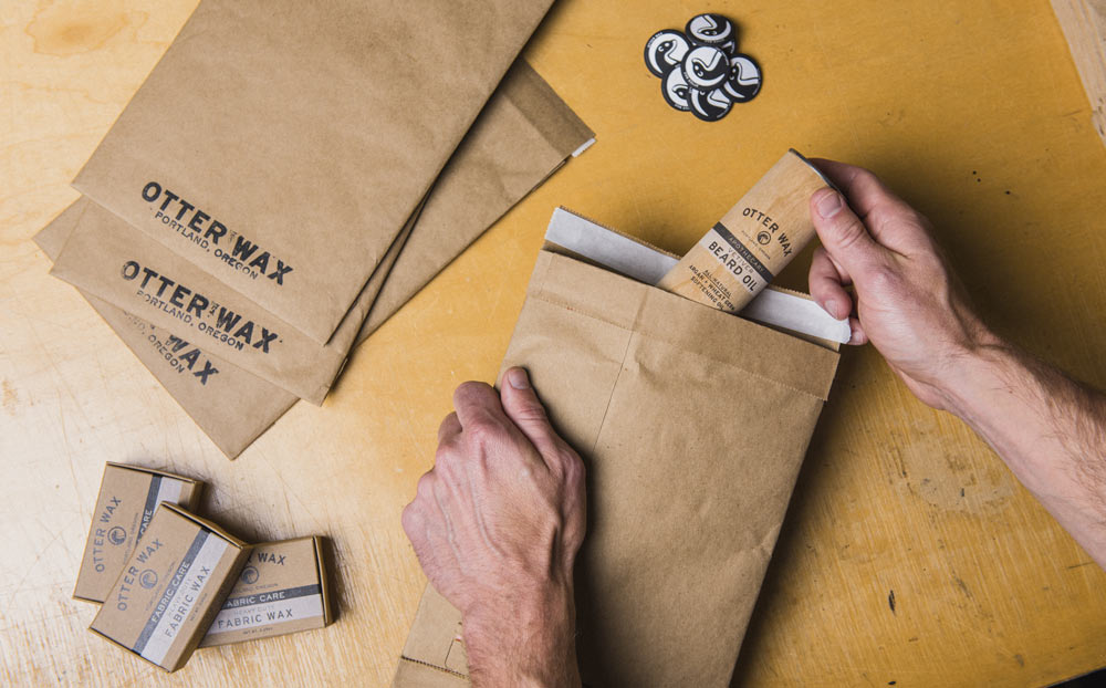 Hands placing an Otter Wax Beard Oil into a brown paper shipping bag surrounded by Fabric Wax bars and Otter Wax–branded envelopes.