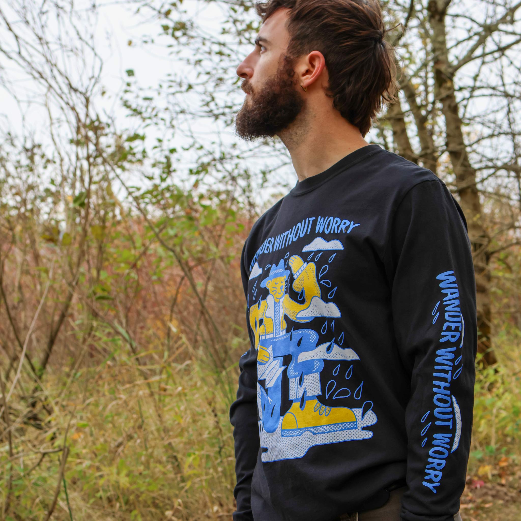Man wearing an Otter Wax brand black long-sleeve shirt with a graphic design and text 'Wander Without Worry' in a forest setting.