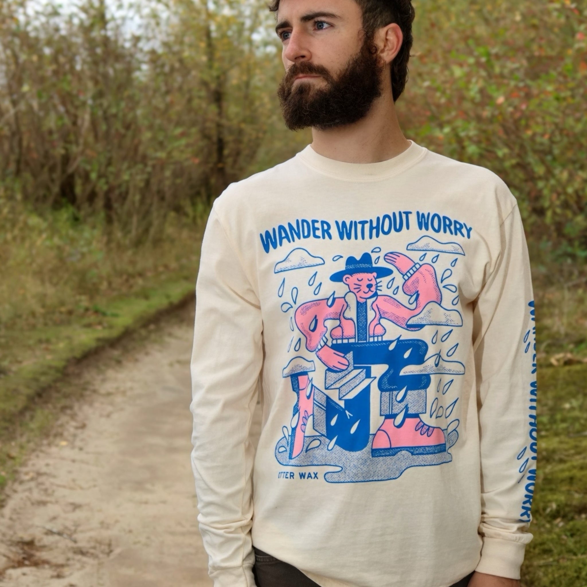 A bearded man standing on a forest path wearing an ivory long-sleeve shirt with a colorful blue and pink illustration and the words “WANDER WITHOUT WORRY” on the front. The shirt features playful cartoon figures and raindrops, and “OTTER WAX” branding near the hem.