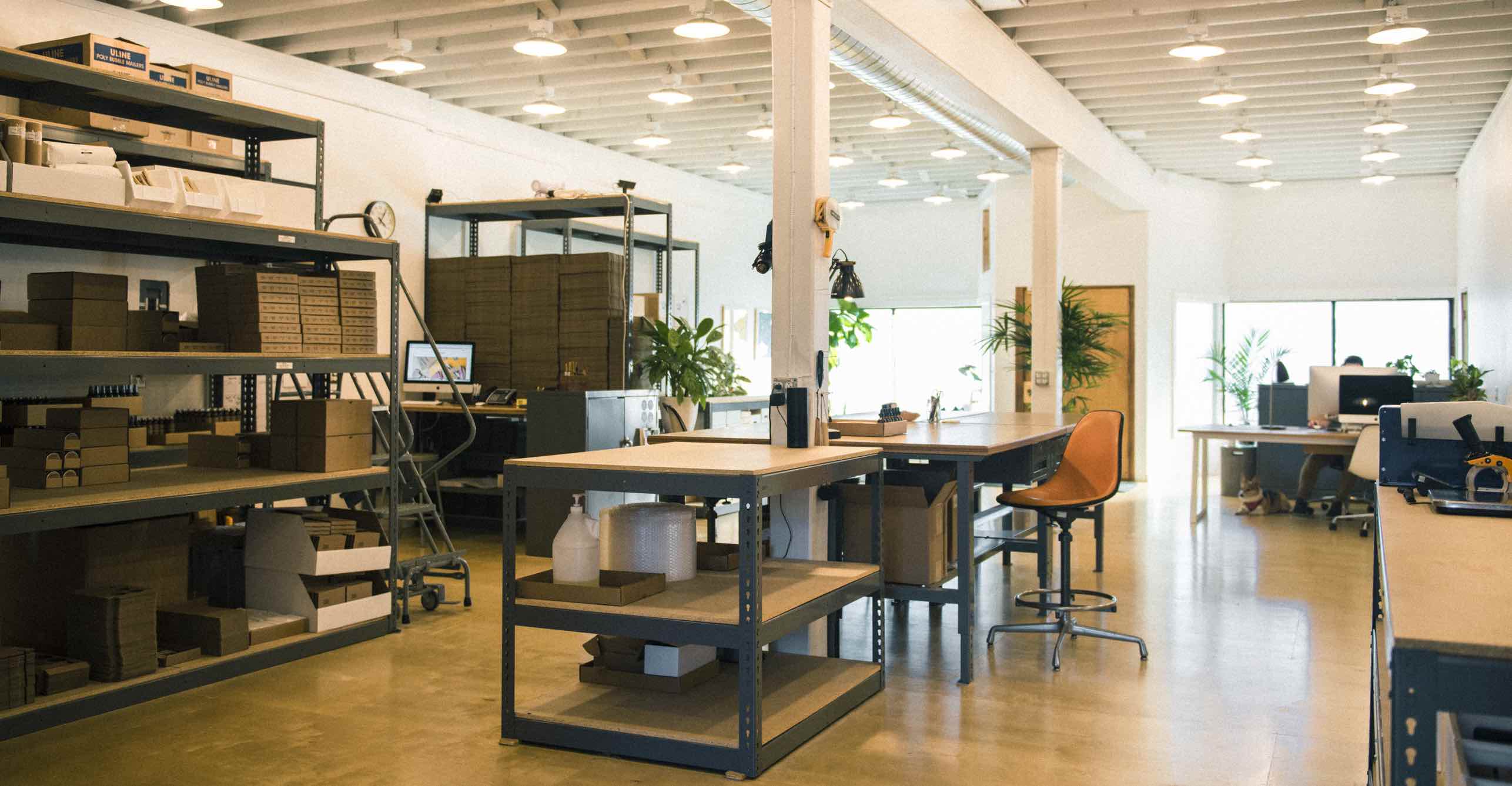 Interior of the Otter Wax workshop and office showing work tables, shelving with packaging supplies, and bright natural light from large windows.